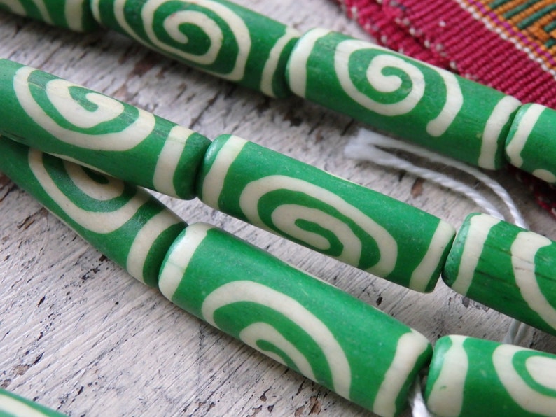 May include: Close-up of green and cream-colored wooden beads with a spiral design. The beads are strung together with white string. The background is a light-colored, textured surface.