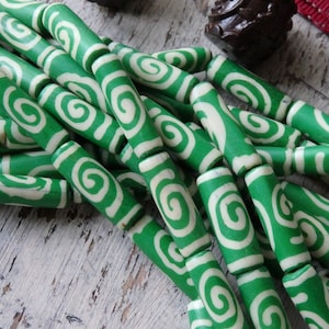 May include: Green and white swirl pattern beads. The beads are cylindrical with a bright green background and a white spiral design. The beads are strung together, suggesting they are for jewelry making or crafts.