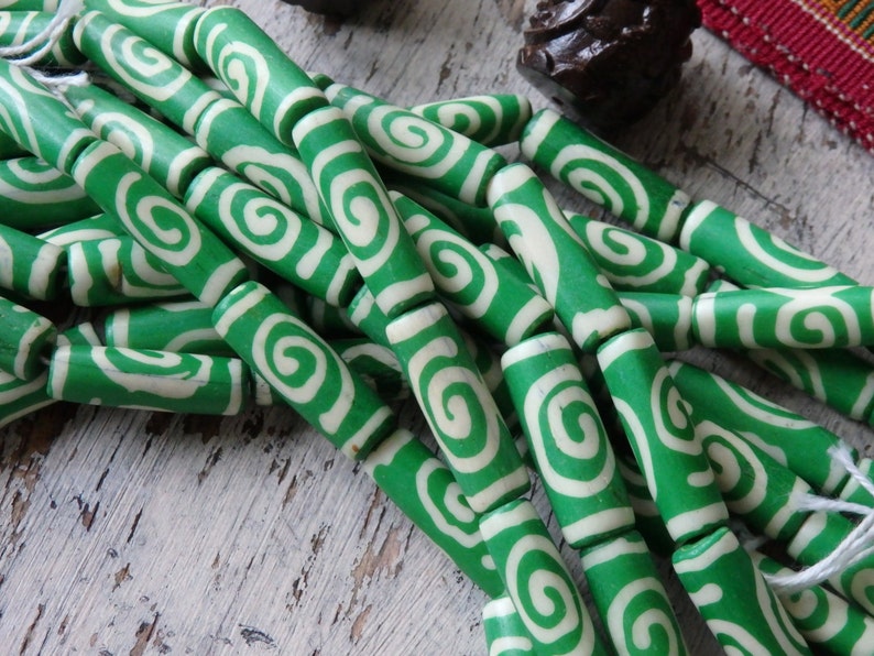 May include: Green and white swirl pattern beads. The beads are cylindrical with a bright green background and a white spiral design. The beads are strung together, suggesting they are for jewelry making or crafts.
