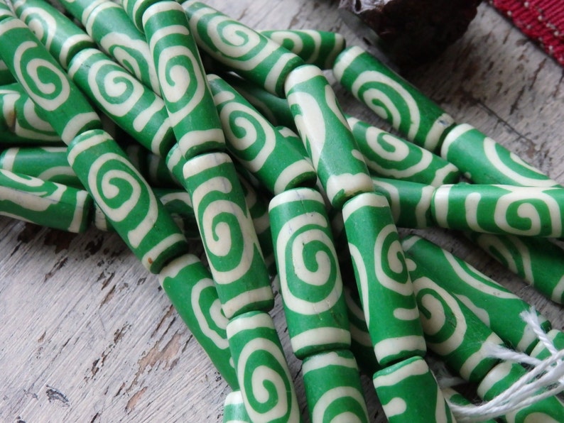 May include: A collection of green and white swirl-patterned beads. The cylindrical beads are a vibrant green with a cream-colored spiral design. The beads are strung together, suggesting they are for jewelry making or crafting.