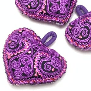 May include: Three heart-shaped ornaments featuring purple and black embroidered paisley patterns. Each heart has a small purple fabric loop attached to the top edge for hanging. The embroidery consists of intricate swirls and lines.