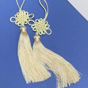 Könnte beinhalten: Zwei hellgelbe chinesische Knoten-Anhänger mit Quasten. Die Knoten sind quadratisch und haben ein gewebtes Design. Die Quasten sind lang und aus einem weichen, cremefarbenen Material gefertigt.