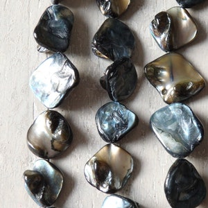 Może przedstawiać: Szereg iryzujących czarnych i niebieskich koralików z muszli. Koraliki mają kwadratowy kształt i naturalny, organiczny wygląd.