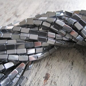 May include: A strand of silver-toned, rectangular, faceted beads. The beads are arranged in a loose pile, showing the reflective surface of the beads.