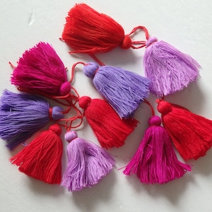 May include: Assortment of colorful tassels in red, pink, and purple. The tassels are made of soft yarn and have a fluffy texture.