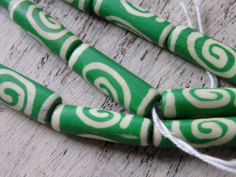 May include: Close-up of green and cream-colored wooden beads with a spiral design. The beads are cylindrical and strung together with white string. The beads are a vibrant green color with a cream-colored spiral pattern.
