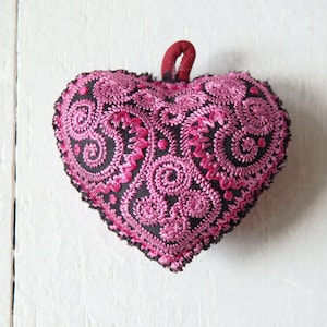 May include: A heart-shaped decorative item with intricate pink embroidery on a black background. The embroidery features swirling patterns. A small red loop is attached at the top. The item is likely handmade.