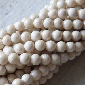 Puede incluir: Una hebra de cuentas redondas de color blanco roto hechas de coral fosilizado. Las cuentas son lisas y tienen una textura natural y orgánica.