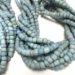 Puede incluir: Múltiples hilos de pequeñas cuentas de vidrio de color azul claro mate. Cada cuenta tiene una forma redondeada y ligeramente irregular con un acabado suave y opaco. Los hilos están agrupados, mostrando el color azul pálido uniforme.
