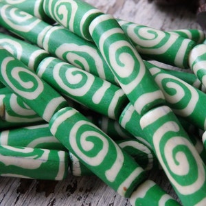 May include: Close-up of green and white swirl beads. The beads are cylindrical with a green exterior and a white, swirled design. The beads are strung together, suggesting they are for jewelry making or crafts.