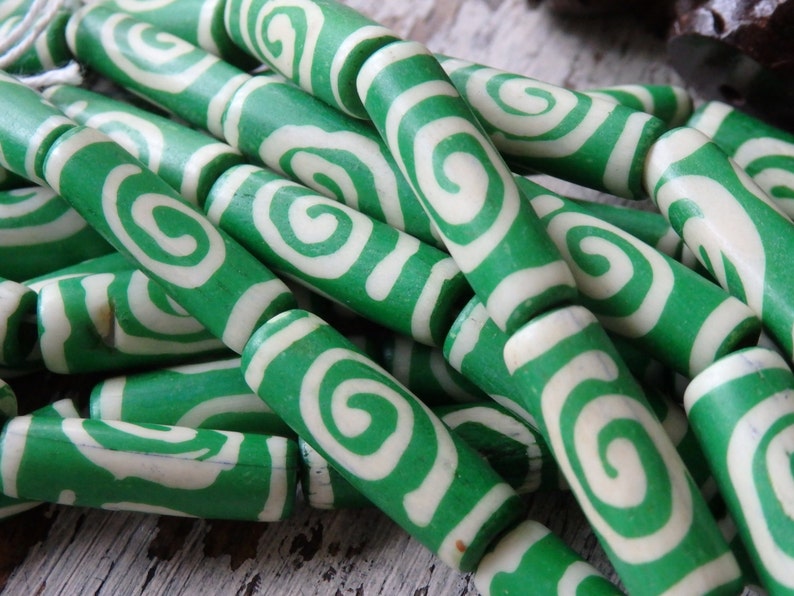 May include: Close-up of green and white swirl beads. The beads are cylindrical with a green exterior and a white, swirled design. The beads are strung together, suggesting they are for jewelry making or crafts.