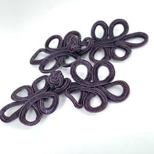 May include: Three purple fabric Chinese knot closures, also known as frog closures, with intricate designs. These decorative closures are often used on clothing and accessories.