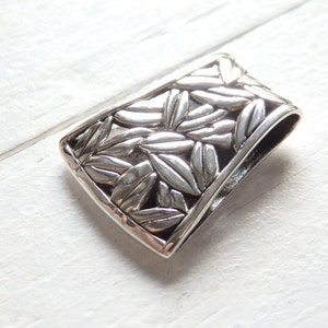 May include: A silver pendant with a detailed leaf pattern. The pendant is rectangular in shape and has a smooth, polished finish.