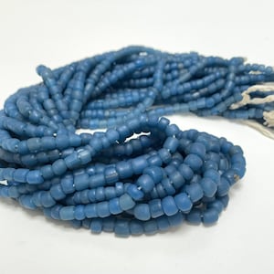 May include: A long strand of small, matte, cornflower blue beads strung together on a white thread. The beads are uniform in size and shape, featuring a slightly irregular, rounded appearance with a soft, non-reflective finish throughout the strand.