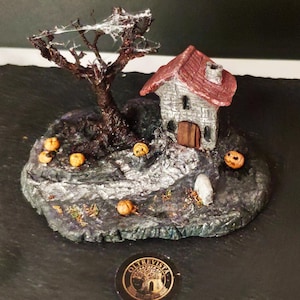 Op de afbeelding: Een miniatuur diorama met een klein stenen huisje met een rood dak, een kale boom met witte spinnenwebben en verschillende oranje pompoenen op een donkere, getextureerde basis. Een kleine zwarte munt met de tekst OLTREVISTA is inbegrepen.