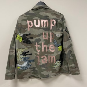 May include: A camouflage jacket with the phrase "pump up the jam" in pink lettering. The jacket has decorative embellishments and is hanging on a metal hanger against a white wall.