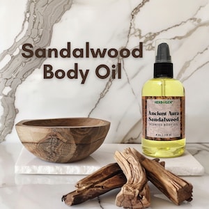 May include: A clear glass bottle of sandalwood scented body oil with a green and white label that reads "HERBOGEN Ancient Aura Sandalwood Scented Body Oil 4 oz | 118 ml". The bottle is sitting on a white marble surface with a wooden bowl and pieces of driftwood.