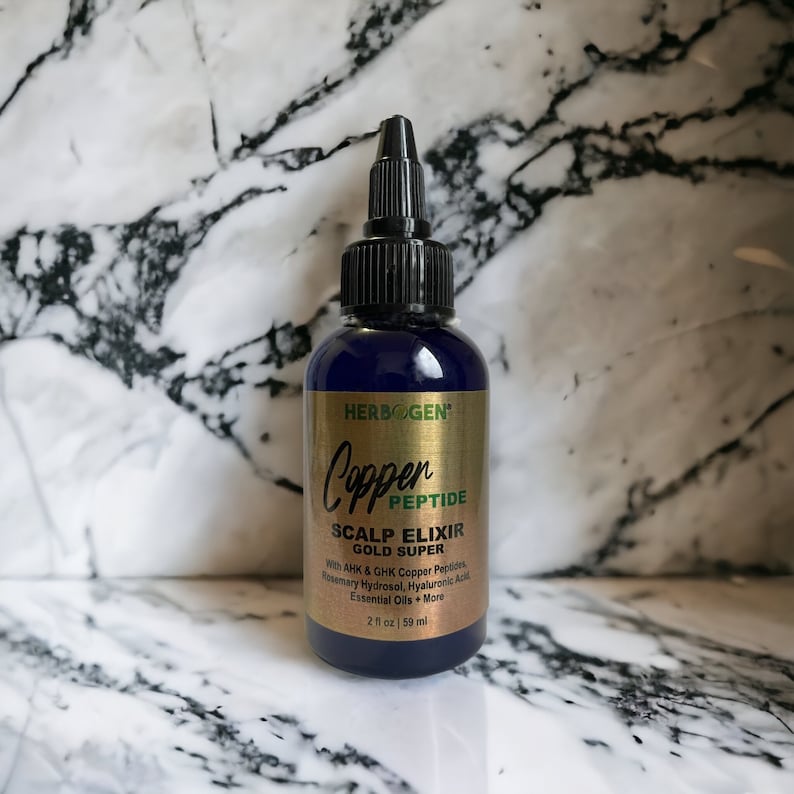 May include: A dark blue plastic bottle of Herbogen Copper Peptide Scalp Elixir Gold Super with a black pointed applicator cap. The gold label features black text listing ingredients including AHK and GHK copper peptides, rosemary hydrosol, hyaluronic acid, and essential oils, with a volume of 2 fl oz.