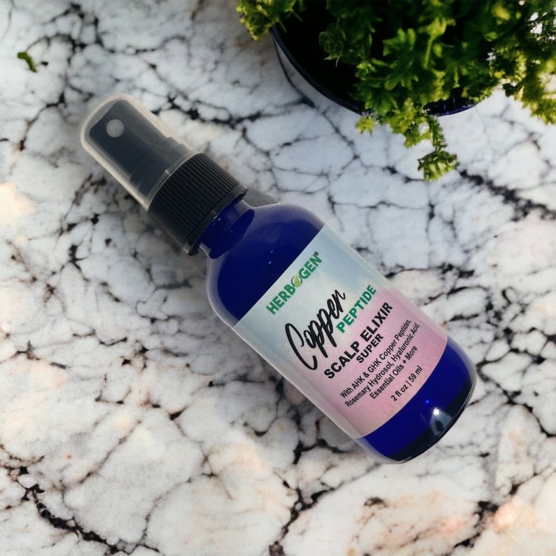 May include: A blue plastic spray bottle of Herbogen Copper Peptide Scalp Elixir. The label features the text Copper Peptide Scalp Elixir Super with AHK & GHK Copper Peptides, Rosemary Hydrosol, Hyaluronic Acid, Essential Oils + More, 2 fl oz | 59 ml.