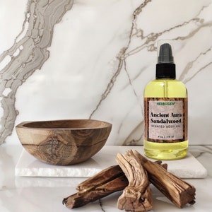 May include: A clear glass bottle of Ancient Aura Sandalwood Scented Body Oil, 4 oz | 118 ml, by HERBOGEN, on a white marble surface with a wooden bowl and pieces of driftwood.