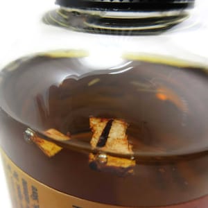 May include: Close-up of a clear glass bottle filled with a dark amber liquid. The liquid contains pieces of dried fruit, possibly orange peel.