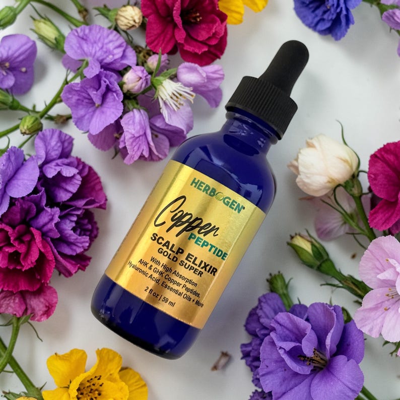 May include: A blue glass bottle with a black dropper cap containing Herbogen Copper Peptide Scalp Elixir Gold Super. The gold label displays the product name, ingredients including AHK and GHK copper peptides, hyaluronic acid, and essential oils, with a volume of 2 fl oz | 59 ml.