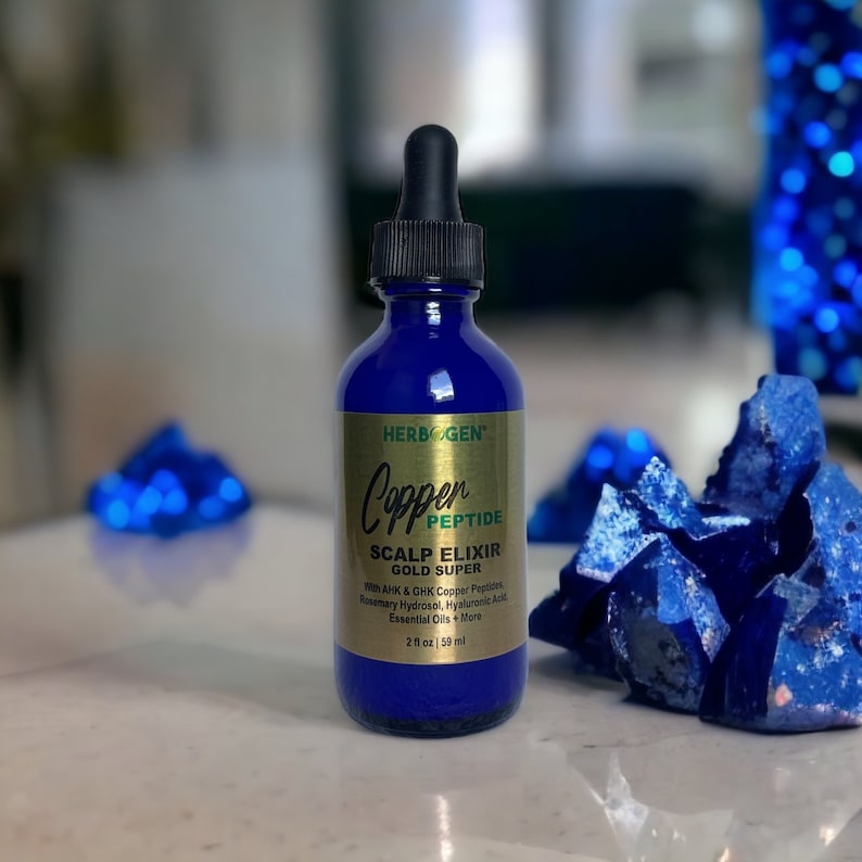 May include: A cobalt blue glass dropper bottle labeled Herbogen Copper Peptide Scalp Elixir Gold Super. The gold label lists ingredients including AHK & GHK Copper Peptides, Rosemary Hydrosol, Hyaluronic Acid, and Essential Oils, with a volume of 2 fl oz.