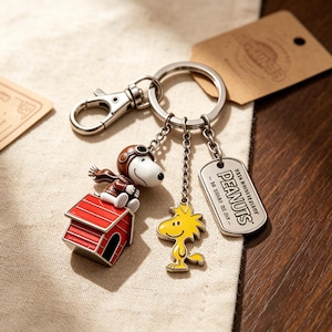 Könnte beinhalten: Ein silberfarbener Metallschlüsselanhänger mit einer Snoopy-Figur, die eine braune Fliegermütze und einen Schal auf einer roten Hundehütte trägt, einem gelben Woodstock-Anhänger und einem rechteckigen Schild mit der Aufschrift PEANUTS.