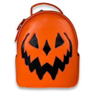 Puede incluir: Una mochila de color naranja brillante que presenta una gran aplicación negra con forma de cara de calabaza dentada en la parte frontal. La bolsa tiene una parte superior redondeada, un asa única y cremalleras plateadas en los laterales.