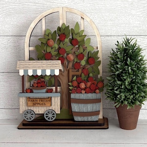 May include: A layered wooden decorative sign featuring an arched window frame, an apple tree with red apples, a small cart labeled FARM FRESH APPLES, and a barrel filled with apples. The sign is displayed next to a small potted green plant.