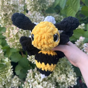 May include: A plush bee toy crafted from soft, chunky yarn featuring a bright yellow head with large black eyes, black wings, and a striped yellow and black body. The stuffed creature has a rounded shape and a textured, fuzzy appearance throughout.