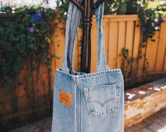 May include: A light blue denim tote bag crafted from repurposed jeans, featuring a prominent tan leather brand patch on the left and a functional back pocket on the right, complete with two sturdy denim shoulder straps.