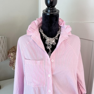 May include: A long-sleeved button-down shirt featuring a pink and white vertical striped pattern, a ruffled collar, and a single chest pocket. The shirt is displayed on a black mannequin and is accessorized with a silver-toned statement necklace.