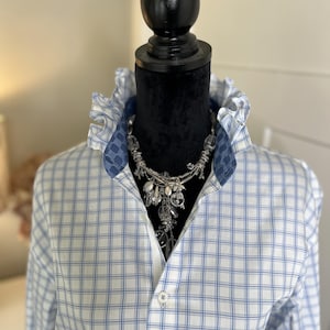 May include: A white button-down shirt with a blue grid pattern, featuring a high ruffled collar with a dark blue interior lining. A multi-strand silver-toned necklace with clear, faceted crystal-like beads hangs around the neck.
