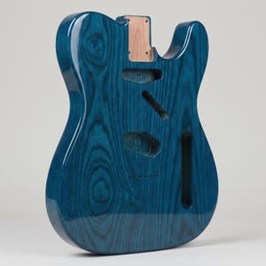 Könnte beinhalten: Ein E-Gitarrenkorpus in glänzendem, durchscheinendem Blau, der eine ausgeprägte Holzmaserung aufweist. Der Korpus verfügt über eine Halstasche, gefräste Aussparungen für Tonabnehmer und Elektronik sowie sechs Löcher für die Saitenführung.