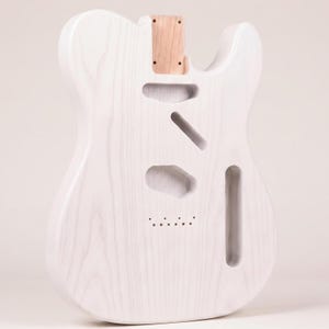 Puede incluir: Un cuerpo de guitarra eléctrica sin terminar hecho de madera de color claro con un patrón de veta visible. El cuerpo presenta un diseño de corte simple, un bolsillo para el mástil y cavidades pre-enrutadas para pastillas, puente y electrónica.