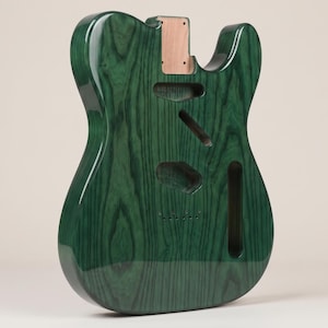 Puede incluir: Un cuerpo de guitarra eléctrica sin terminar con un acabado en verde esmeralda profundo y un patrón de veta de madera visible. El cuerpo presenta un bolsillo para el mástil, cavidad para el puente y ranuras de control, con madera natural.