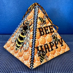 May include: A pyramid-shaped needlepoint pillow with a bee and honeycomb design. The pillow is decorated with the words "Bee Happy" and features multiple bees.