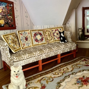 Könnte beinhalten: Ein Miniatur-Wohnzimmer mit einer Couch, Kissen, einem Teppich, einem weißen Hund und einer schwarzen Katze. Die Couch ist mit einem floralen Stoff bezogen und hat fünf Kissen mit gestickten Designs. Der Teppich ist ebenfalls gemustert und hat einen hellblauen Hintergrund. Der Hund sitzt auf dem Teppich und die Katze sitzt auf einem Regal im Hintergrund.