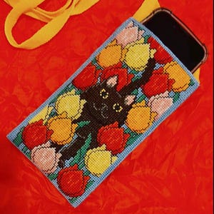 Puede incluir: Funda de teléfono bordada con un gato negro rodeado de tulipanes rojos, amarillos y rosas. La funda tiene un borde azul. Una correa amarilla es visible sobre un fondo rojo.