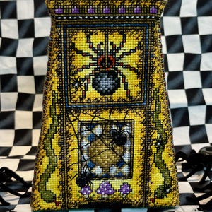 Może przedstawiać: Poduszka haftowana krzyżykiem w kolorze żółtym i zielonym z wzorem pajęczyny i wieloma pająkami. Poduszka ma kształt piramidy i ma purpurowo-złotą lamówkę.