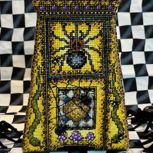 Może przedstawiać: Poduszka haftowana krzyżykiem w kolorze żółtym i zielonym z wzorem pajęczyny i wieloma pająkami. Poduszka ma kształt piramidy i ma purpurowo-złotą lamówkę.