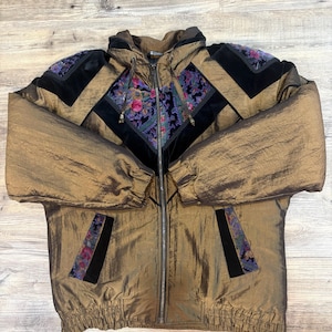 May include: A metallic bronze hooded jacket featuring black velvet panels and floral patterned fabric accents on the chest, shoulders, and pockets. The jacket includes a full-length front zipper, long sleeves with elastic cuffs, and a ribbed hem.