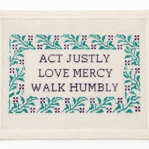 May include: A rectangular cross-stitch embroidery on cream-colored fabric featuring the text ACT JUSTLY, LOVE MERCY, WALK HUMBLY in dark purple thread. The text is surrounded by a decorative border of teal leaves and small purple floral accents.