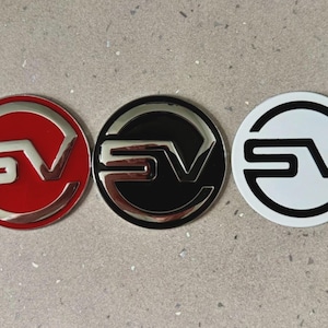 Könnte beinhalten: Drei kreisförmige Embleme mit den Buchstaben SV in einer stilisierten Schriftart. Das erste Emblem hat einen roten Hintergrund mit silberner Schrift, das zweite einen schwarzen Hintergrund mit silberner Schrift und das dritte einen weißen Hintergrund mit schwarzer Schrift.