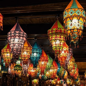 May include: A collection of multi-tiered fabric lanterns hanging from a dark ceiling. Each lantern features vibrant colors including orange, pink, blue, green, and yellow, with intricate geometric patterns, diamond shapes, and decorative tassels.