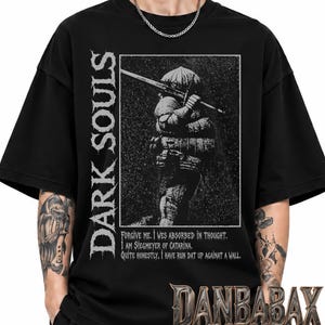 Könnte beinhalten: Ein schwarzes Kurzarm-T-Shirt mit einer weißen Grafik eines Ritters in abgerundeter Rüstung, der ein Schwert hält. Der Text lautet vertikal "DARK SOULS" und "Forgive me. I was absorbed in thought. I am Siegmeyer of Catarina. Quite honestly, I have run dat up against a wall."