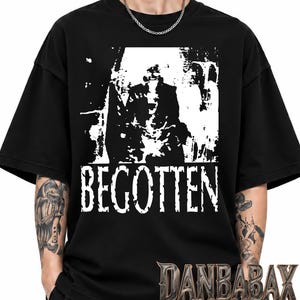 Op de afbeelding: Een zwart T-shirt met korte mouwen en een contrastrijke witte grafische print van een figuur in een masker en gewaden. Het woord BEGOTTEN is in een verweerd wit lettertype onder de afbeelding gedrukt. Het shirt heeft een ronde hals.