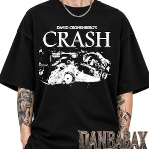 Op de afbeelding: Een zwart T-shirt met korte mouwen en een witte grafische print van een autowrak. De tekst boven de afbeelding luidt DAVID CRONENBERG'S CRASH in een vetgedrukt wit lettertype. Het shirt heeft een ronde hals en een losse, boxy pasvorm.