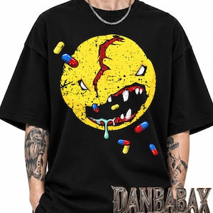 Może przedstawiać: Czarny t-shirt z krótkim rękawem z dużą, postarzaną żółtą grafiką przedstawiającą stworzenie z ostrymi zębami, pękniętą głową i śliną. Projekt zawiera unoszące się czerwone i niebieskie kapsułki oraz napis DANBABAX wykonany metaliczną czcionką.